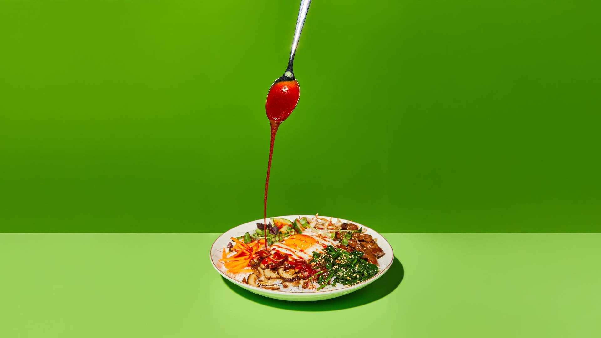 Una salsa rossa viene versata con un cucchiaio su un piatto colorato con molteplici ingredienti condito con un uovo fritto.