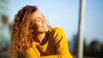 Donna con maglione arancione, in piedi in un prato fiorito con cielo azzurro e muretto a secco sullo sfondo.