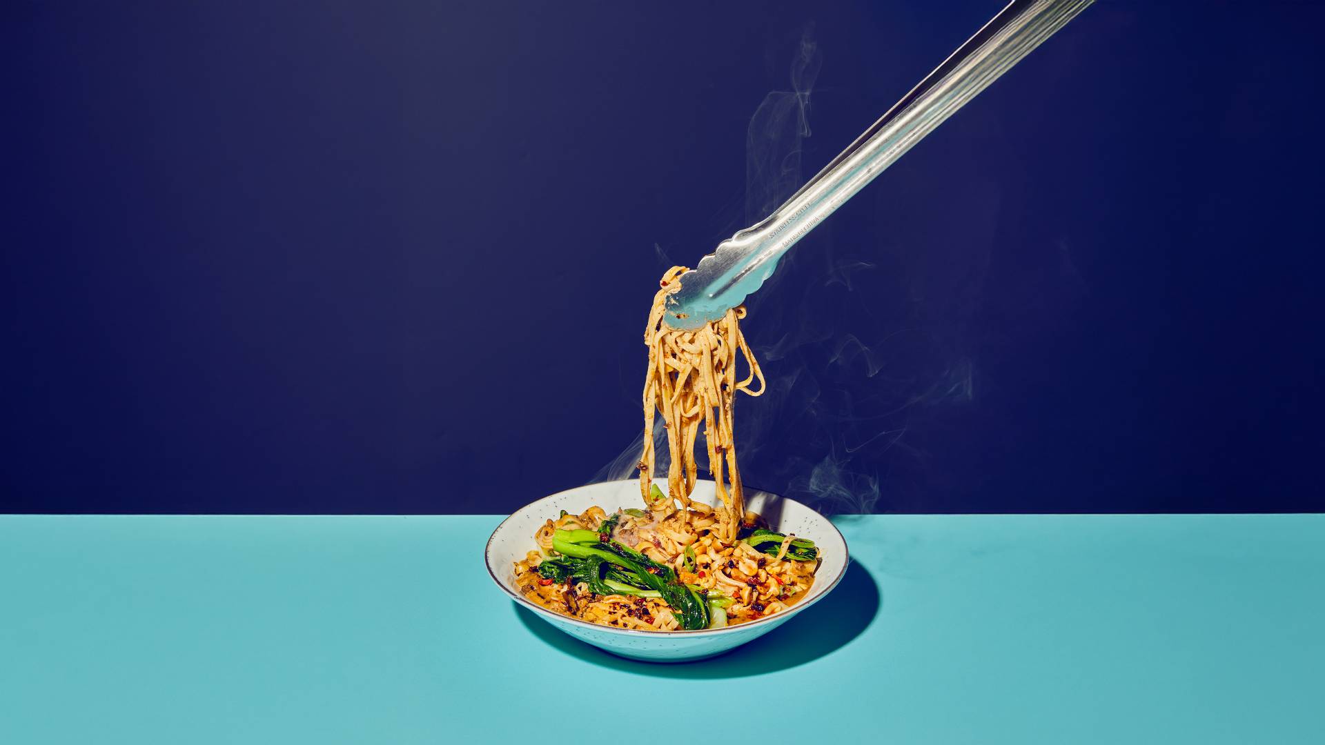 Un'immagine di spaghetti asiatici che vengono sollevati con delle bacchette da una ciotola bianca, fotografate su uno sfondo diviso in blu scuro e chiaro.