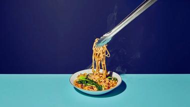 Un'immagine di spaghetti asiatici che vengono sollevati con delle bacchette da una ciotola bianca, fotografate su uno sfondo diviso in blu scuro e chiaro.