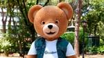 Mascotte orso Coccolino con T-shirt del marchio e gilet di jeans in un parco verdeggiante