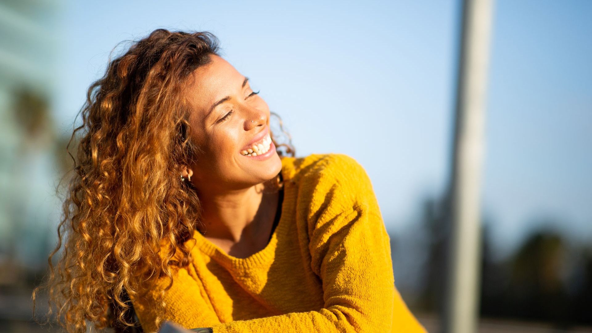 Donna con maglione arancione, in piedi in un prato fiorito con cielo azzurro e muretto a secco sullo sfondo.