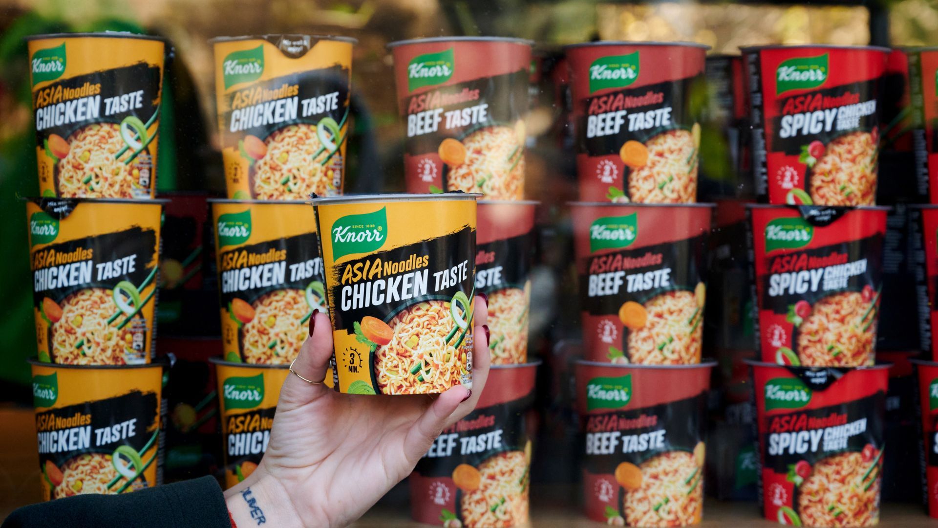 Una mano tiene in mano un vasetto di noodles al pollo su uno sfondo di 16 vasetti misti al gusto di pollo, carne e piccante